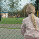 En ung jente står på utsiden av et gjerde og ser inn på en basketballbane der andre ungdommer spiller
