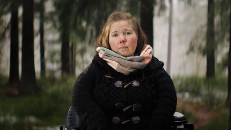 En kvinne i rullestol ser i kamera. Hun er ute i skogen og har på seg sorte klær og et lyst skjerf.