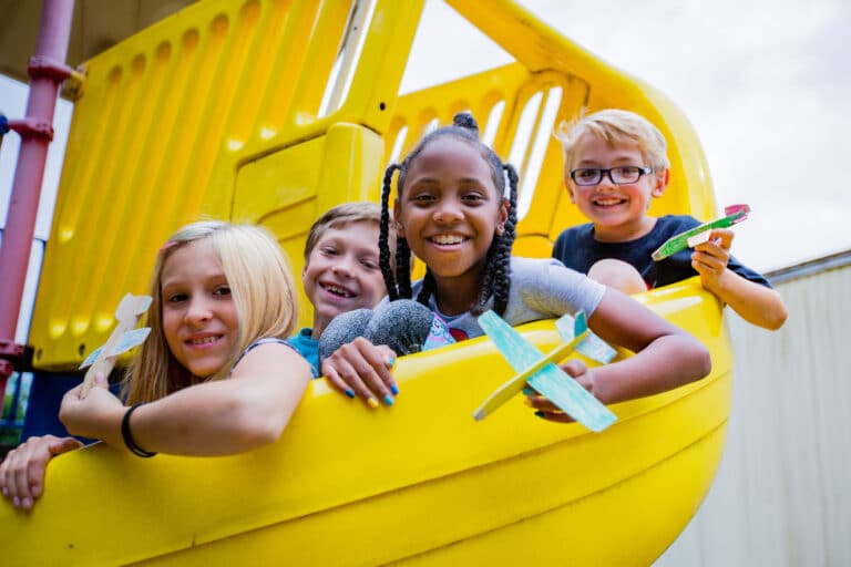 Fire barn sitter med papirfly i hendene på en gul sklie i et klatrestativ. De ser i kamera og smiler.