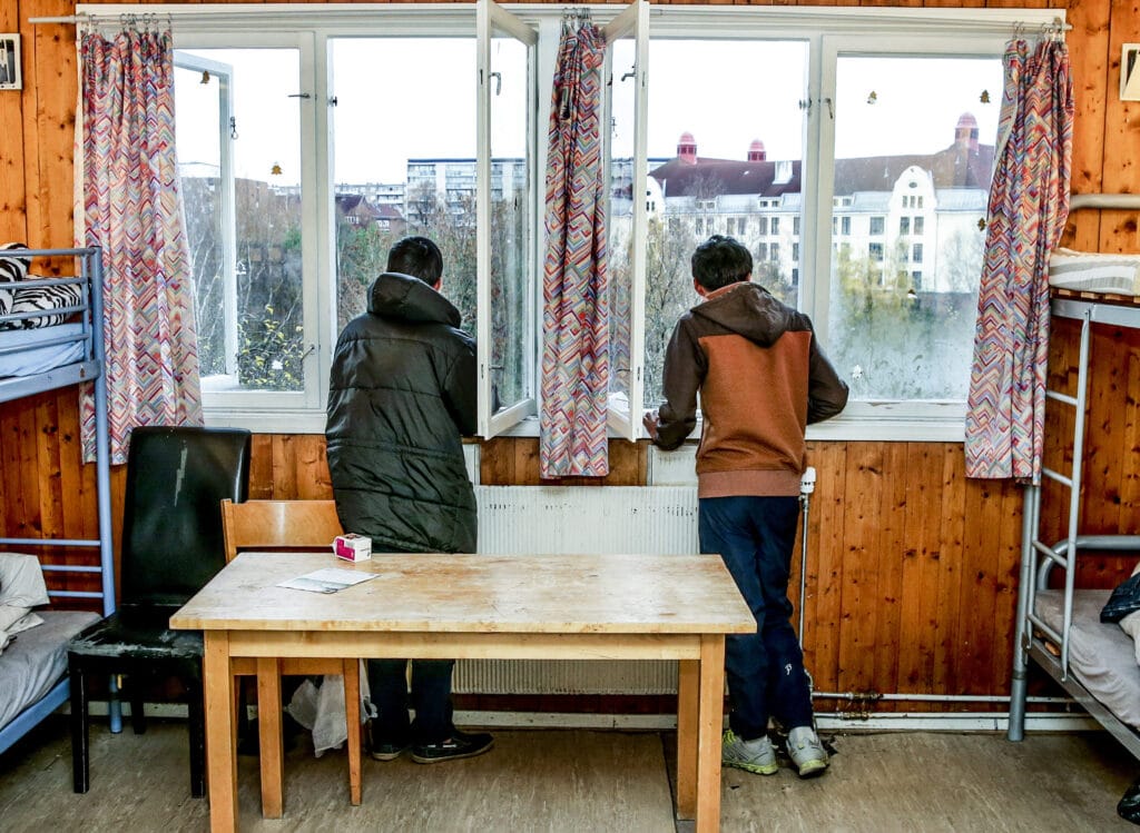To unge gutter med mørkt hår står i et sliten rom med ryggen til og ser ut av vinduet.