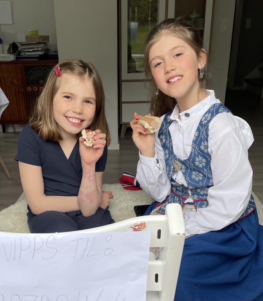 To små jenter som smiler og holder pølse med brød i hver sin hånd. De er pyntet i penttøy og de feirer 17.mai.