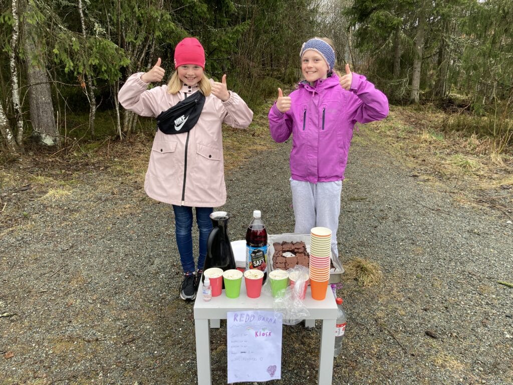 To jenter holder tomlene sine opp. De stårforan et bord som har kopper, en saftflaske, kaffekanne og kake.