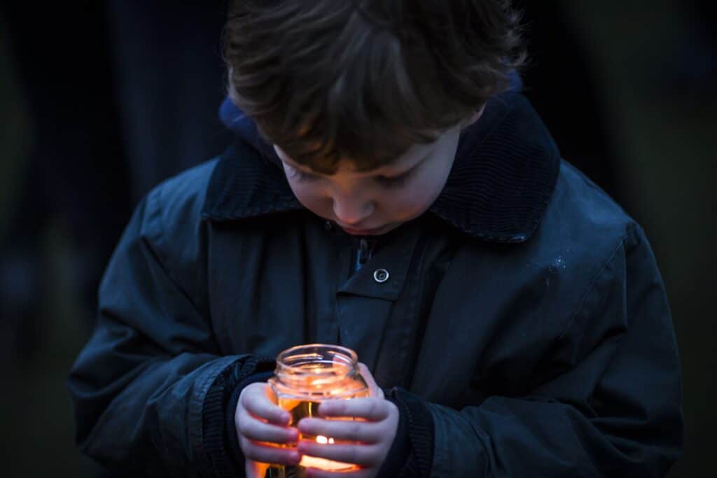 Barn i blå jakke holder et glass med et tent lys mellom hendene, og ser ned på flammen. Få tips til hvordan du kan snakke med barn om 22. juli