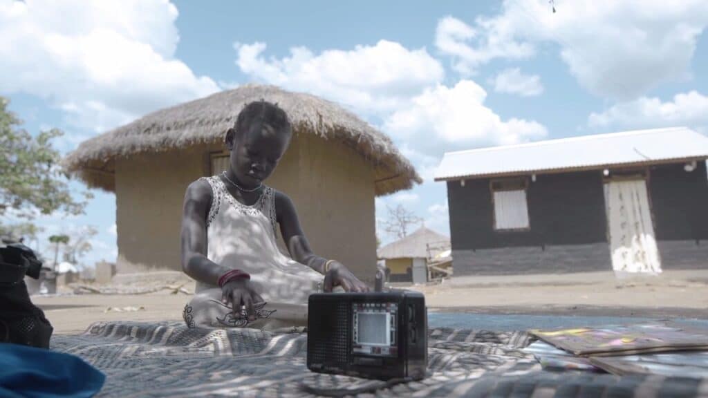 En 6 år gammel jente i hvit kjole sitter på et teppe på bakken utenfor hjemmet sitt i Uganda og har hjemmeundervsining via en liten radio. Fra Redd Barnas rapport om utdanning