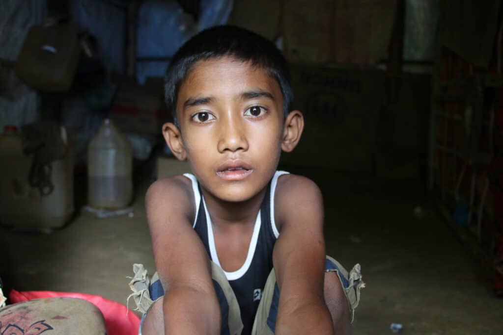 En gutt i en flyktningeleir i Bangladesh sitter på gulvet i hjemmet sitt og kikker inn i kamera. Hjemmet et rammet av klimakrisen, med stadige oversvømmelser og jordskred.