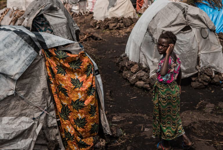Liliane, en 13 år gammel jente står i fordrivelsesleiren hun og familien måtte dra til på grunn av flom. Hun står ved siden av flere telt og mye gjørme.