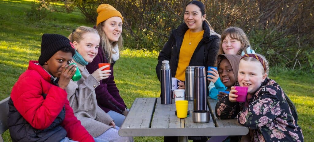 Voksne og barn sitter sammen rundt et piknikbord og drikker varm drikke fra termos