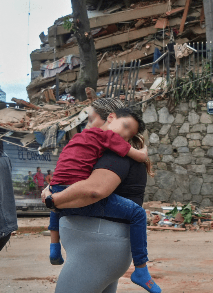 En kvinne bærer et barn foran en totalskadd bygning i Venezuela