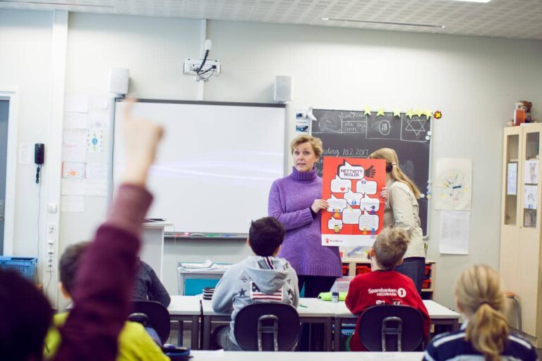Bilde av en lærer i et klasserom som holder opp en plakat med Nettvettregler. I forkant av bildet ser vi bakhodene på elever, og en hånd som rekkes i været.