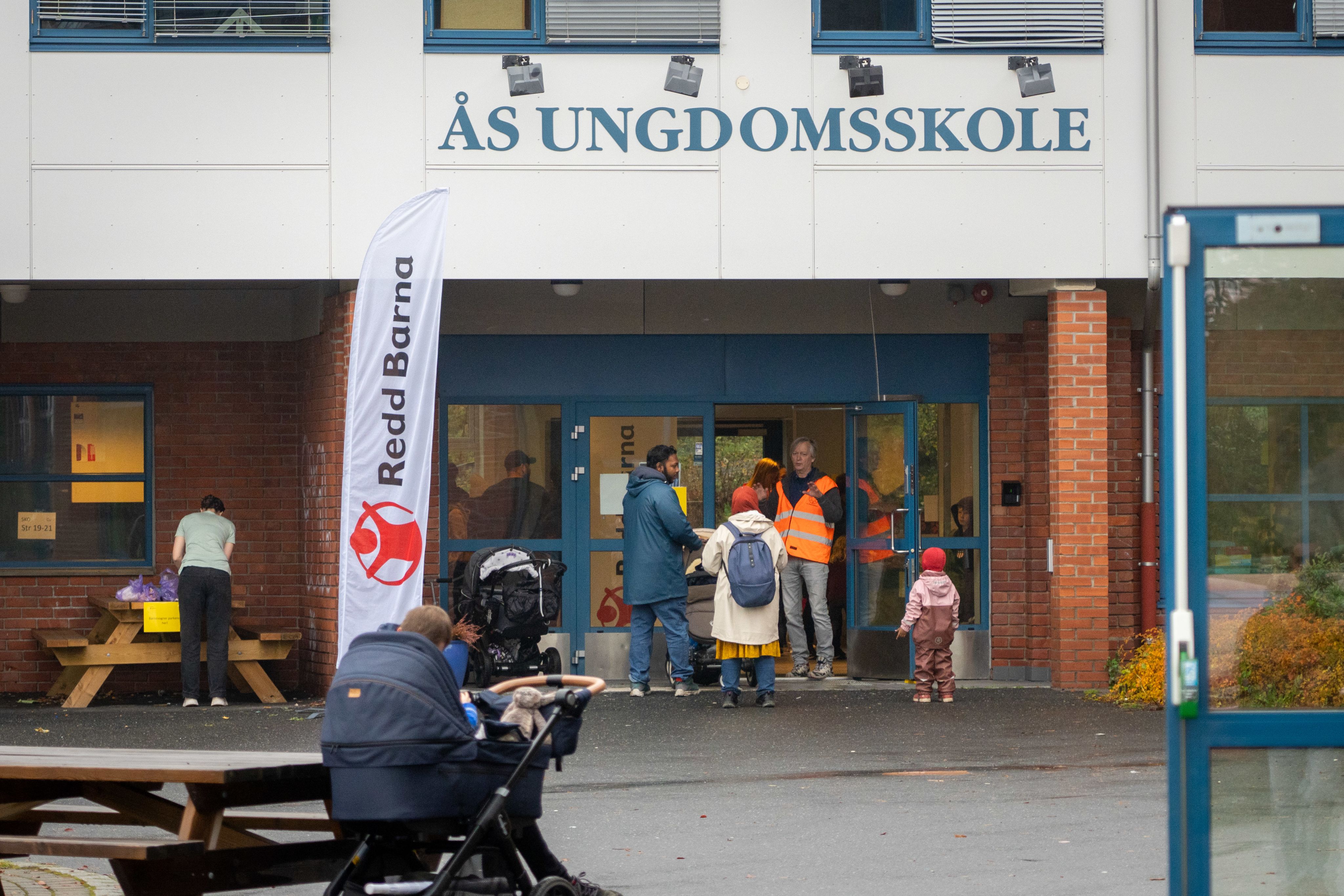 Et Redd Barna banner er plassert ved siden av inngangen til Ås ungdomskole hvor folk er på vei inn.
