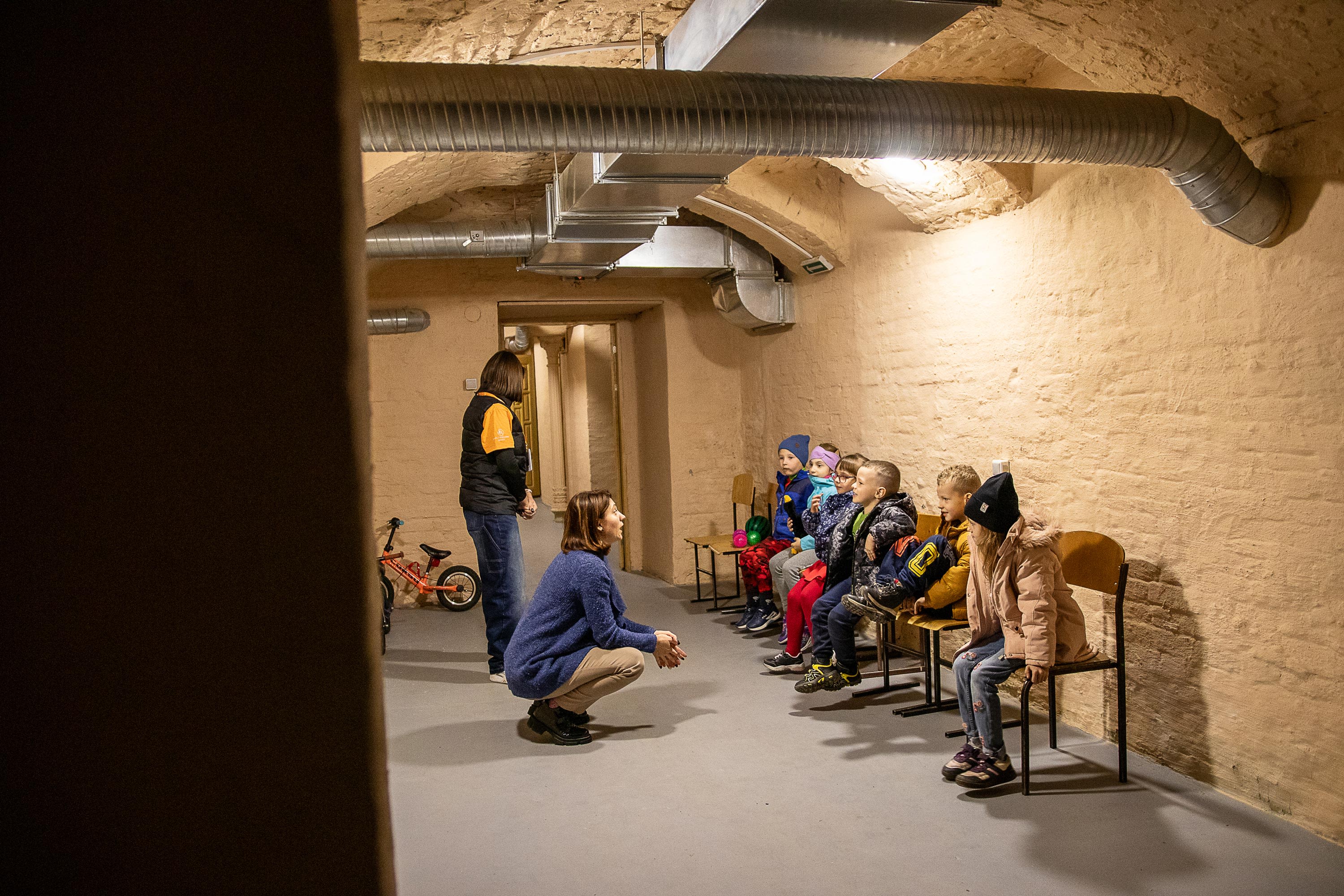 En dame sitter på huk og snakker til barna som sitter langs veggen i kjellerrommet.