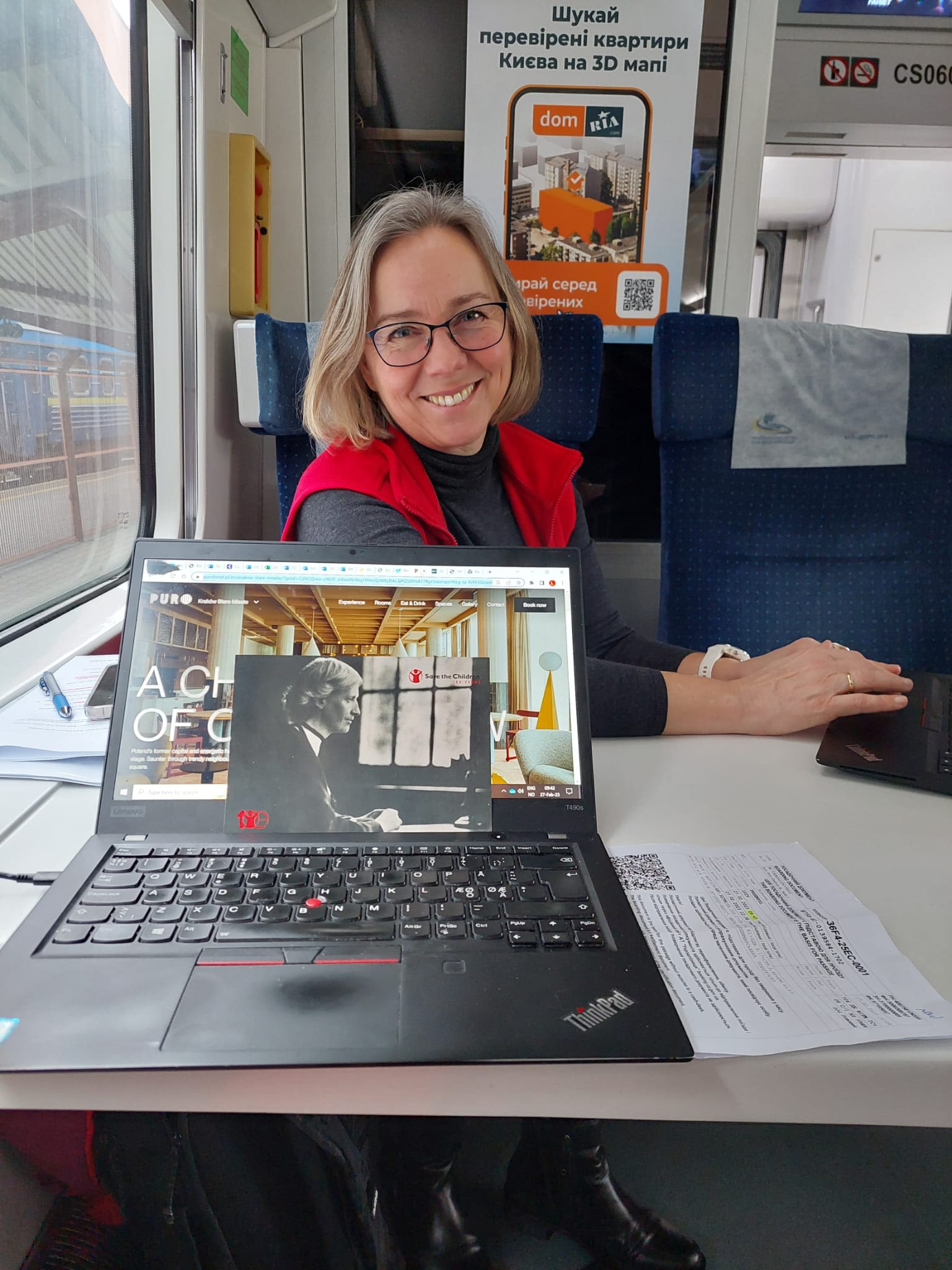 En kvinne i et togsete ser på kamera og smiler. En åpen pc står på bordet foran henne.