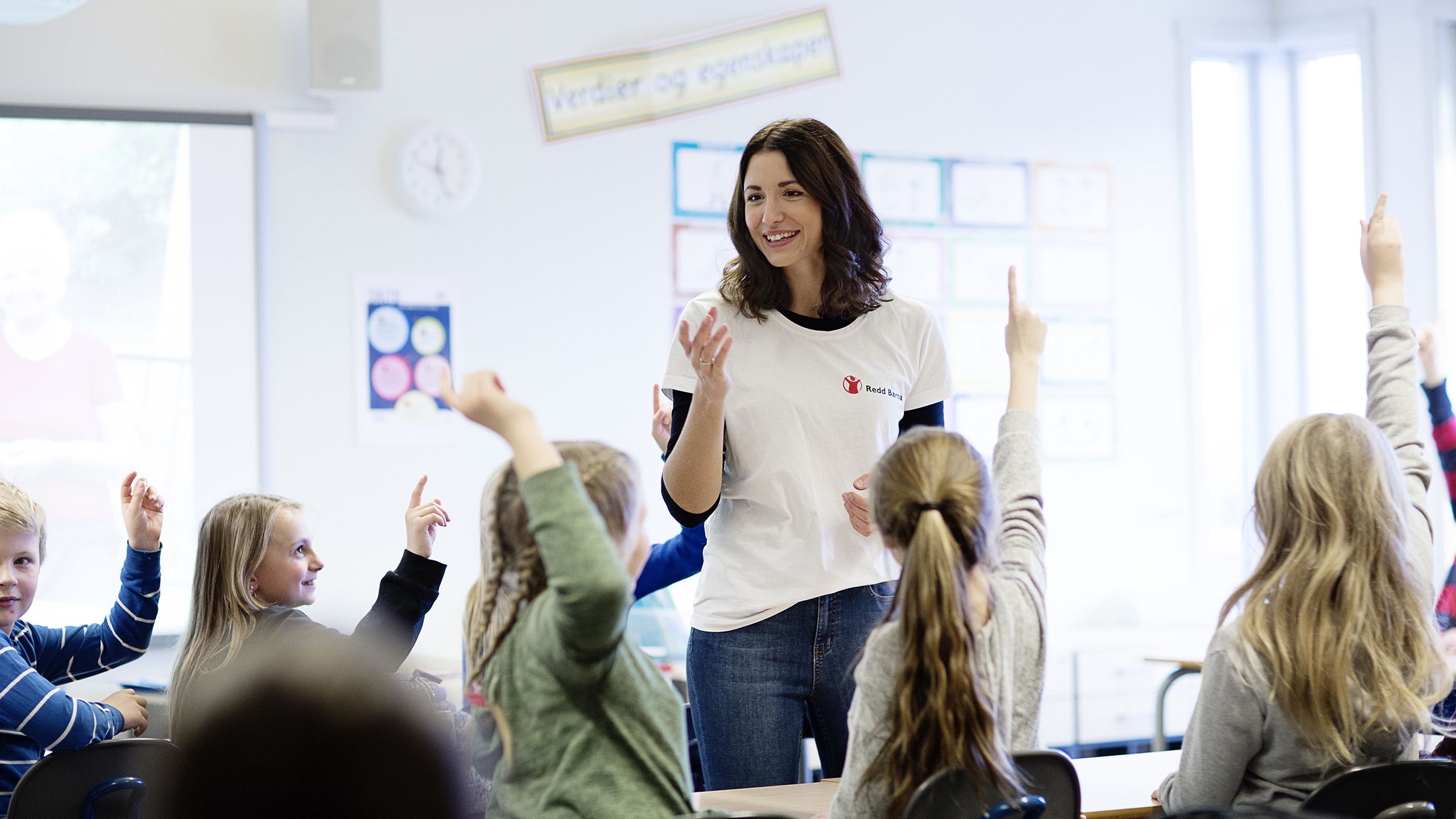En ung kvinne står midt i et klasserom der barn rundt henne rekker opp hånden.