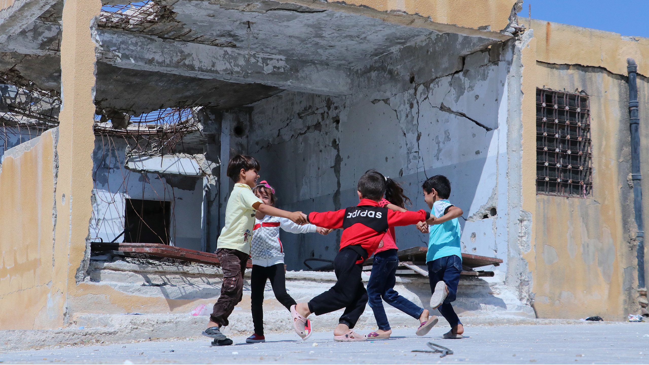 Barn som holder hender og danser i ring foran en utbombet skole.